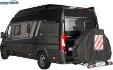 Schwarzer Kastenwagen mit Hochdach und Seitenscheiben, Heckansicht von rechts; montiertes Reserverad mit dunkler Abdeckung und roter-weißer Diagonalwarntafel; Firmenlogo "HINDERMANN" oben links