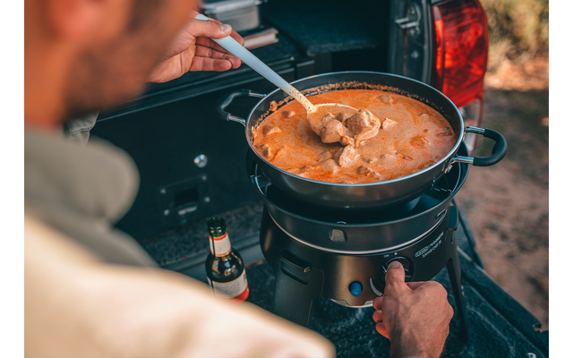 Personne remue avec une cuillère blanche dans une marmite noire contenant un ragoût orange avec des morceaux de viande sur un réchaud de camping portable à l'arrière d'un véhicule; bouteille de bière à côté