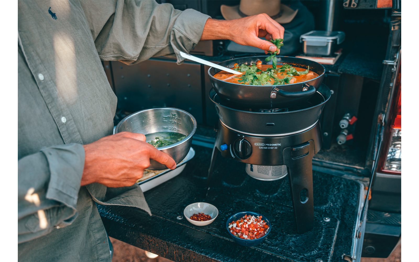 Mains et haut du corps d'une personne en chemise à manches longues ajoutant des herbes fraîches dans une casserole posée sur un réchaud portable ; petits bols avec épices et ingrédients hachés à côté
