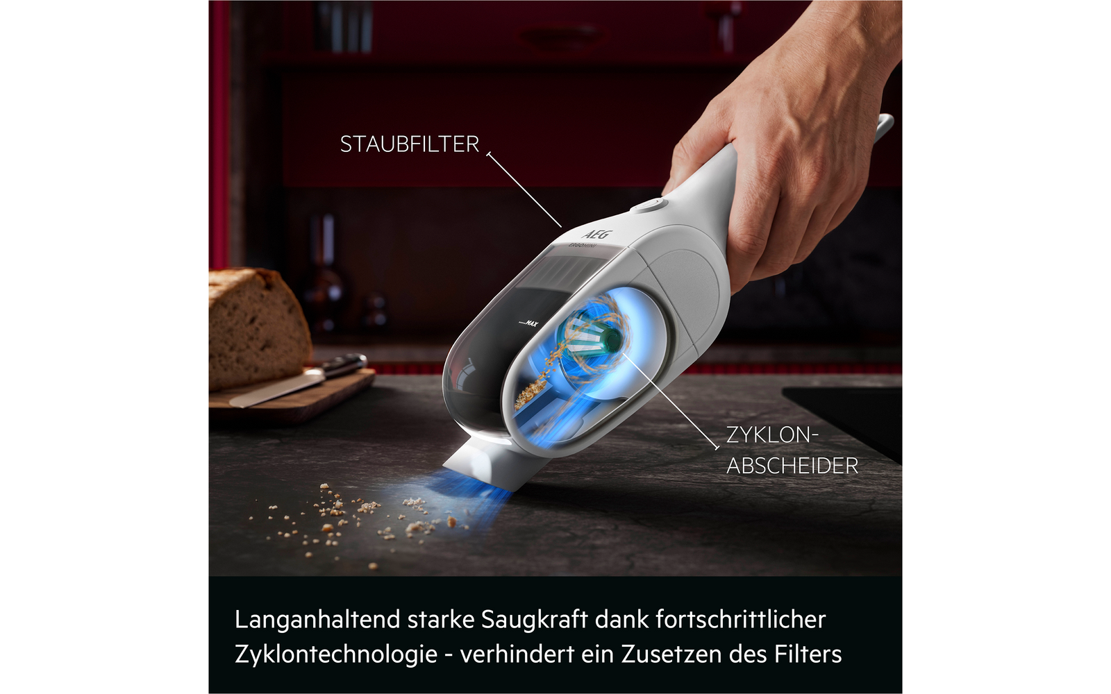 Hand hält weißen Handstaubsauger über dunkler Arbeitsfläche mit Krümeln; durchsichtiges Gehäuse zeigt blauen Wirbel; Beschriftungen 'STAUBFILTER' und 'ZYKLON-ABSCHIEDER'; Brot auf Schneidebrett im Hintergrund; deutscher Werbetext am unteren Bildrand