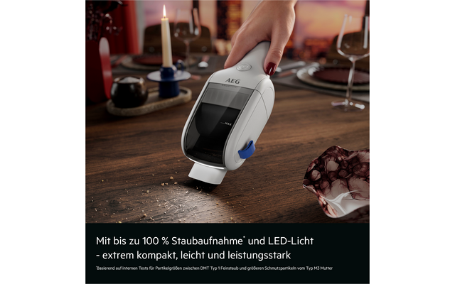 Hand mit rotem Nagellack hält ein weißes AEG-Handstaubsauger über einem Holztisch und saugt Krümel; gedeckter Tisch mit Kerze, Tellern, Weingläsern und Schale im Hintergrund; Textleiste unten mit deutschem Text