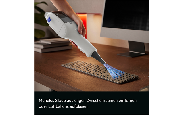 Hand hält weißen Handstaubsauger über schwarze Tastatur auf Holzschreibtisch, blauer Luftstrom grafisch vom Mundstück; im unteren Bildbereich deutscher Text 'Mühelos Staub aus engen Zwischenräumen entfernen oder Luftballons aufblasen'