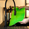 Mani che tengono una spugna verde sotto un getto d'acqua da un rubinetto da cucina nero sopra un lavello nero; sullo sfondo un portaposate
