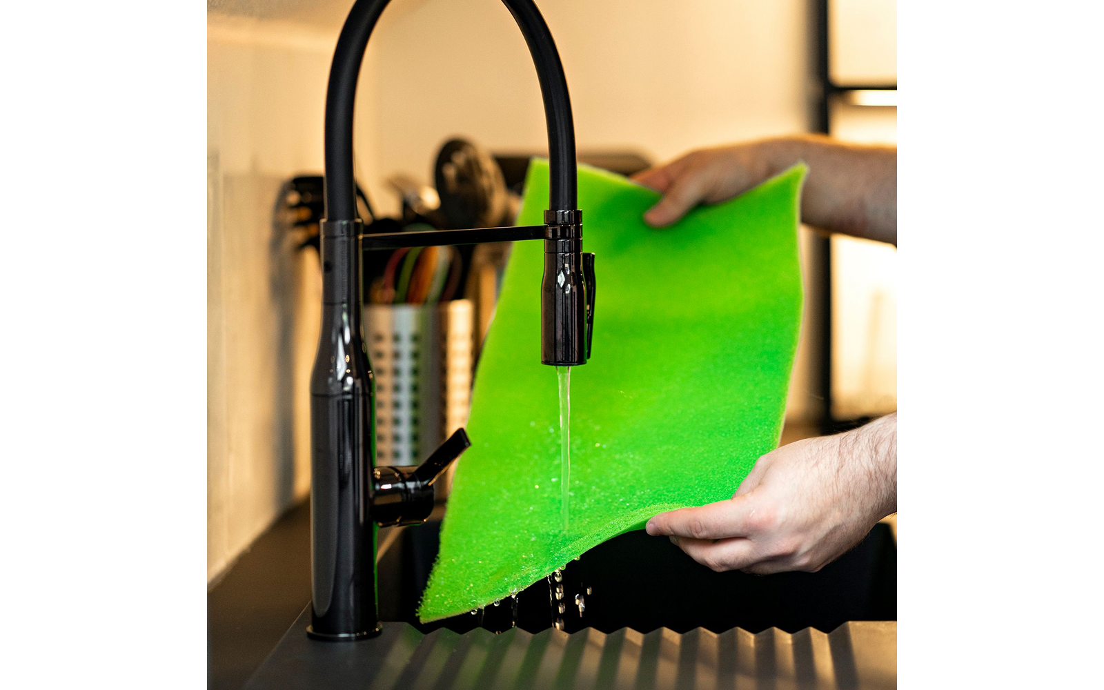 Mani che tengono una spugna verde sotto un getto d'acqua da un rubinetto da cucina nero sopra un lavello nero; sullo sfondo un portaposate
