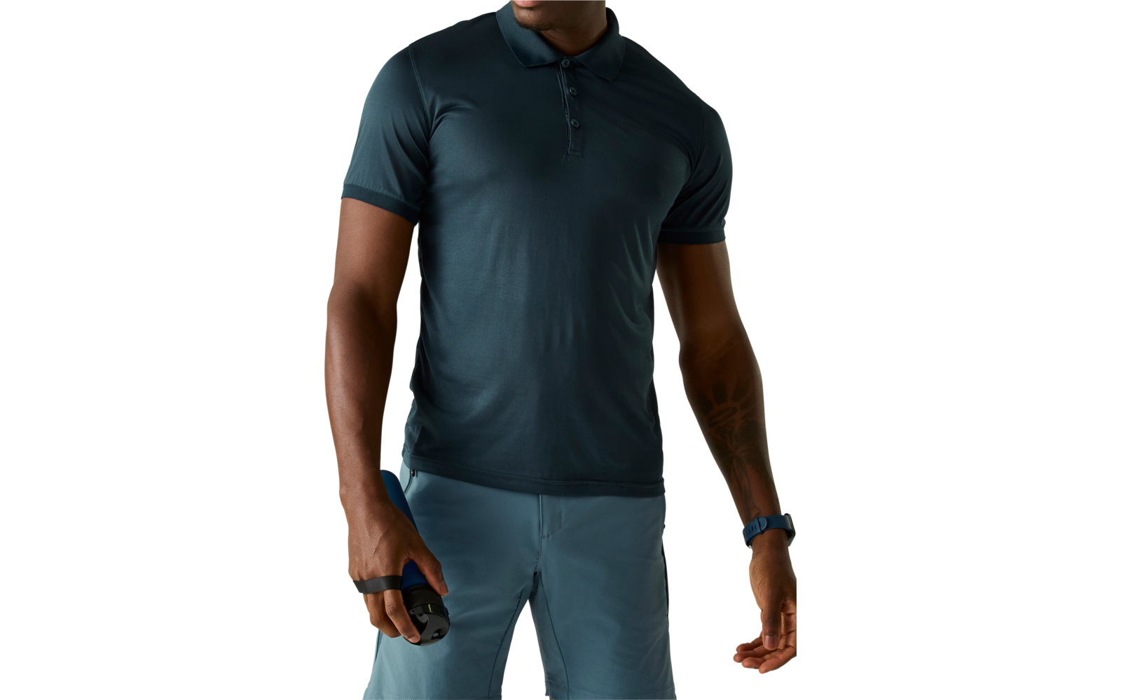 Oberkörper einer Person mit dunkelgrünem Poloshirt und hellblauen Shorts. Linke Hand hält ein kleines schwarzes Gerät. Tätowierung am Unterarm, Gesicht nicht sichtbar