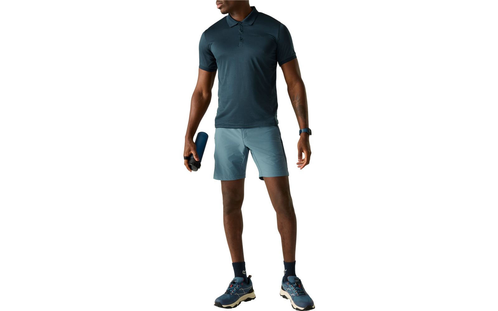 Person in dunkeltürkisfarbenem Poloshirt und hellblauen Shorts, hält eine blaue Flasche, trägt dunkle Socken und Sportschuhe