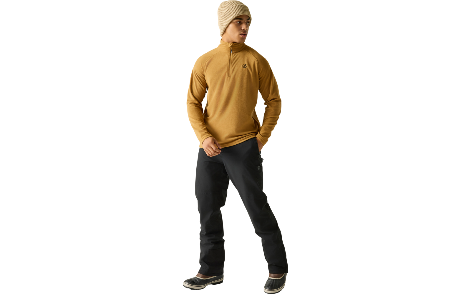 Staande persoon in mosterdkleurige trui met korte rits, beige gebreide muts, zwarte broek en zwarte winterlaarzen met lichte zolen, kijkt opzij