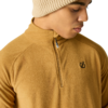 Persoon met beige gebreide muts en mosterdgele fleece met korte rits en klein zwart logo op de linkerborst, kijkt omlaag naar rechts, lichte achtergrond