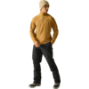 Staande persoon in mosterdkleurige trui met korte rits, beige gebreide muts, zwarte broek en zwarte winterlaarzen met lichte zolen, kijkt opzij