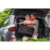 Homme portant des lunettes de soleil tient un jeune enfant blond dans le coffre ouvert d'une voiture argentée et porte un sac de voyage noir avec des détails rouges