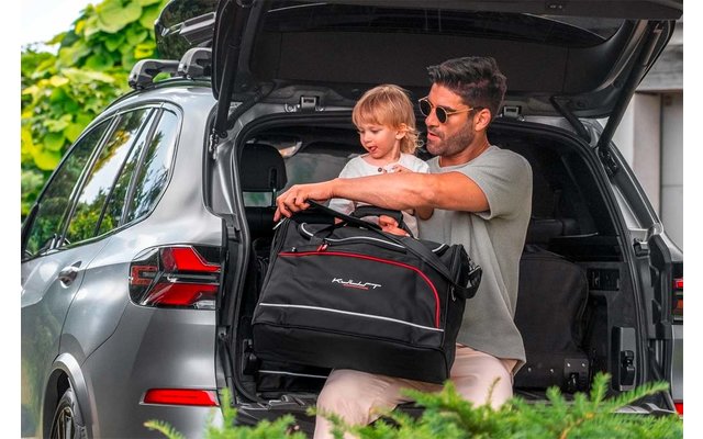 Homme portant des lunettes de soleil tient un jeune enfant blond dans le coffre ouvert d'une voiture argentée et porte un sac de voyage noir avec des détails rouges