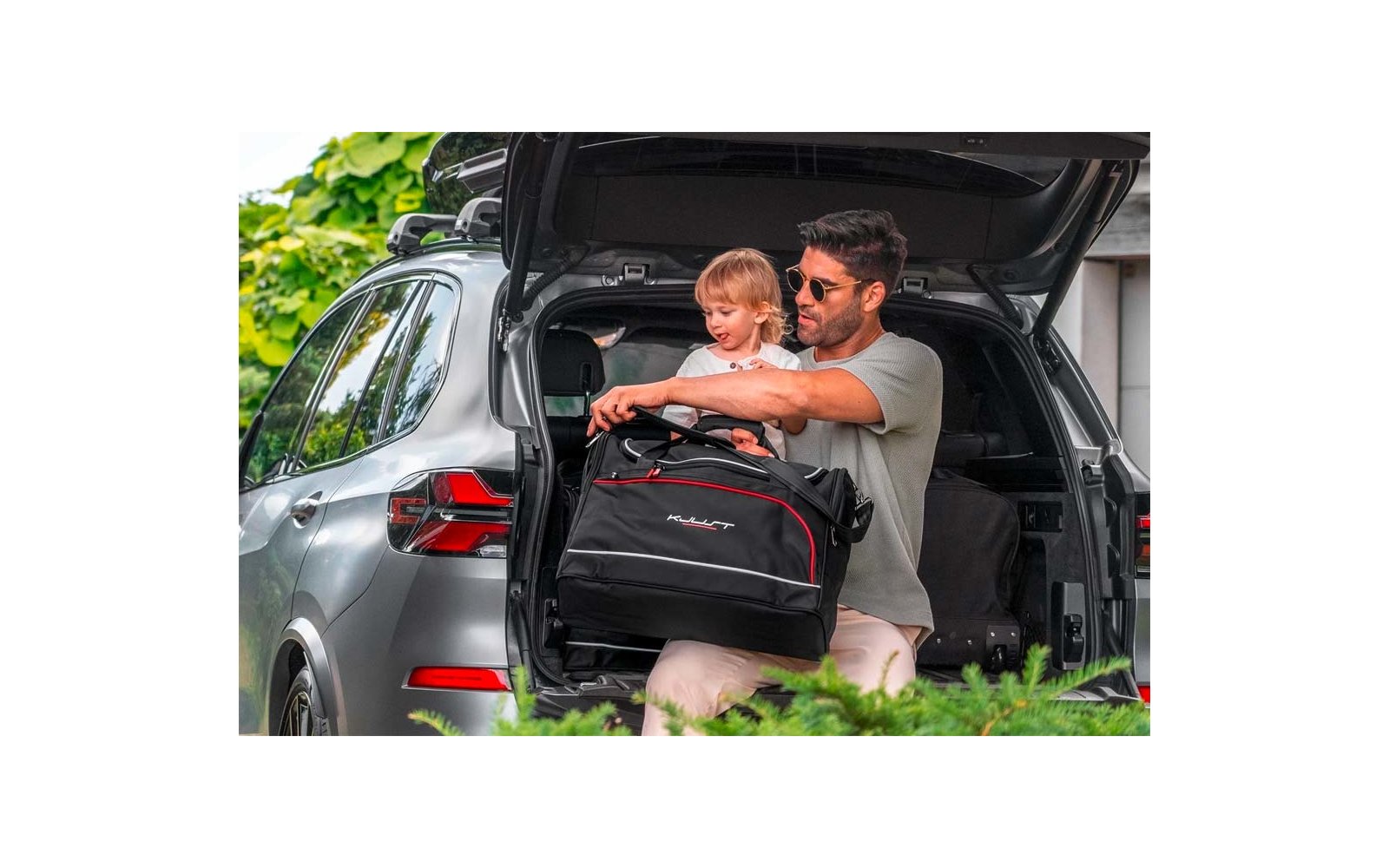 Homme portant des lunettes de soleil tient un jeune enfant blond dans le coffre ouvert d'une voiture argentée et porte un sac de voyage noir avec des détails rouges