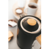 Cafetera negra con jarra de vidrio sobre encimera de madera, junto a tazas de café y cucharas