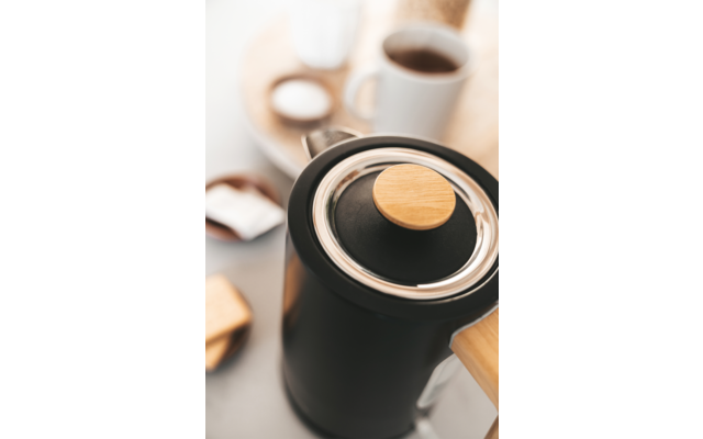 Cafetera negra con jarra de vidrio sobre encimera de madera, junto a tazas de café y cucharas