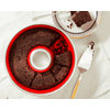 Dark brown round cake in a red ring-shaped pan with one slice removed; the slice on a plate with a fork and crumbs, a cake server beside it on a white tablecloth