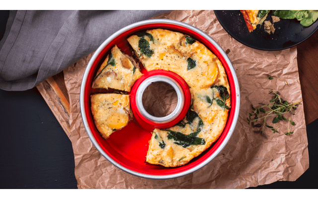 Round red baking pan with golden-brown, pie-slice portions containing green leaf pieces; on crumpled brown paper, gray cloth at left, black plate with one slice at top right, herb sprig at right