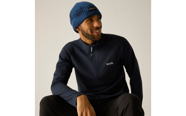 Sitzender Mann mit blauer Strickmütze und dunkelblauem Pullover mit kurzem Reißverschluss, lächelnd und zur Seite schauend