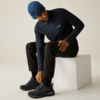 Mann sitzt auf einem weißen Würfel, beugt sich vor und fasst einen Schuh; er trägt eine blaue Strickmütze, einen dunkelblauen Pullover mit Reißverschluss, schwarze Hose und dunkelblaue Schuhe; heller einfarbiger Hintergrund