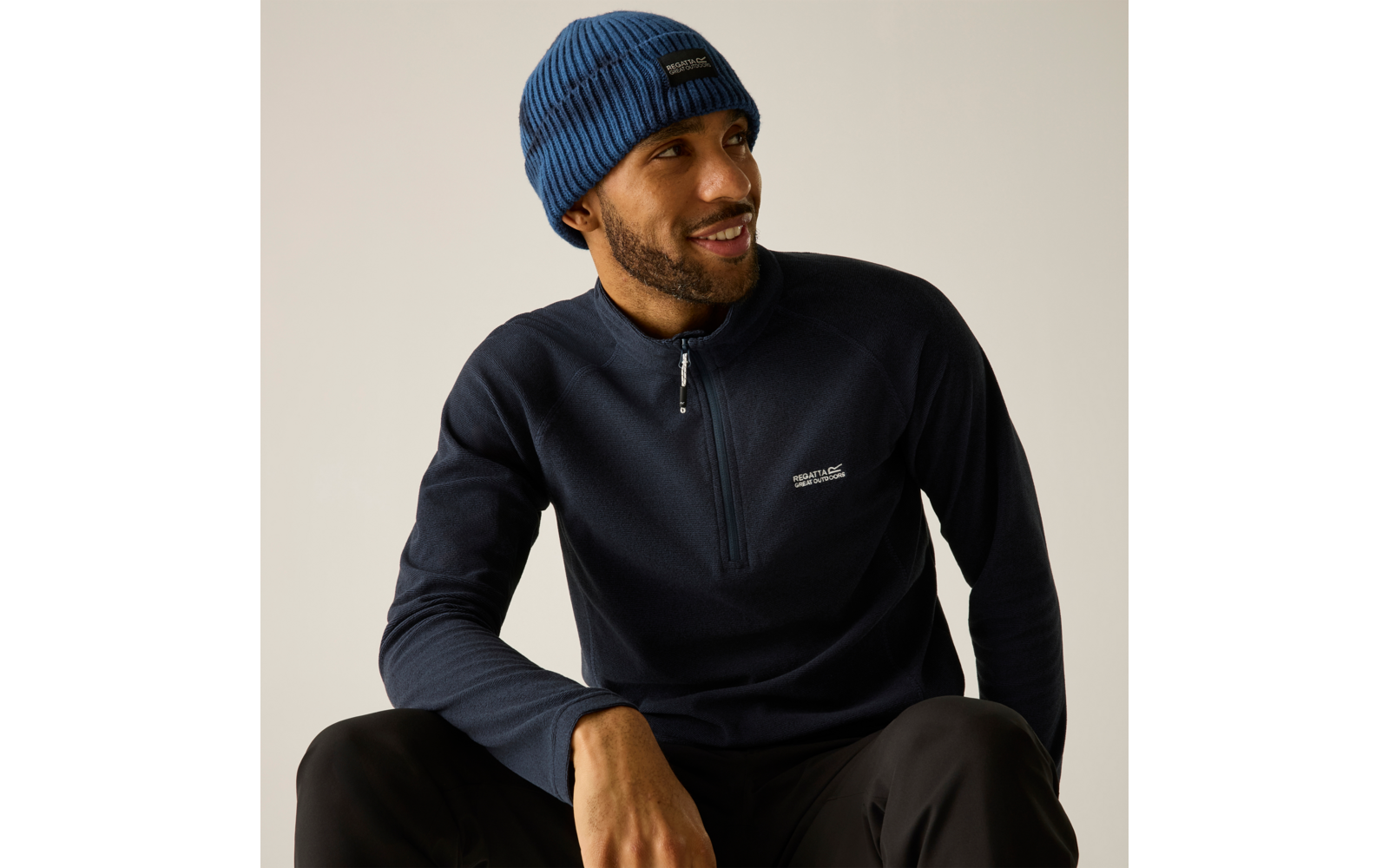 Sitzender Mann mit blauer Strickmütze und dunkelblauem Pullover mit kurzem Reißverschluss, lächelnd und zur Seite schauend