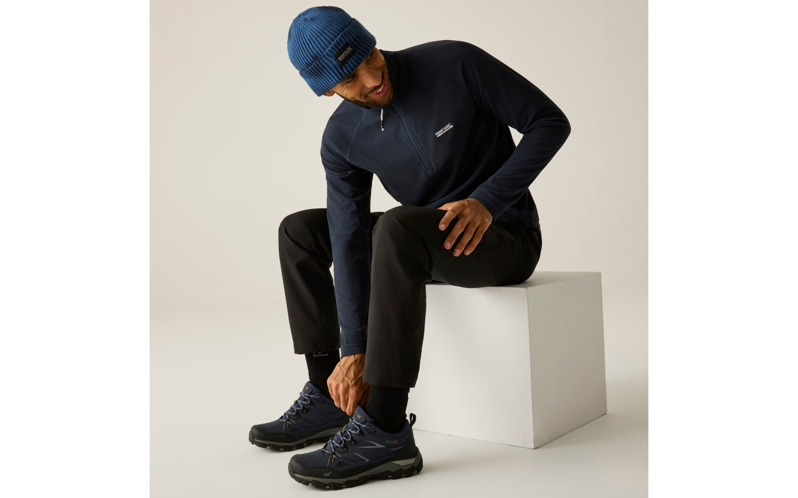 Mann sitzt auf einem weißen Würfel, beugt sich vor und fasst einen Schuh; er trägt eine blaue Strickmütze, einen dunkelblauen Pullover mit Reißverschluss, schwarze Hose und dunkelblaue Schuhe; heller einfarbiger Hintergrund