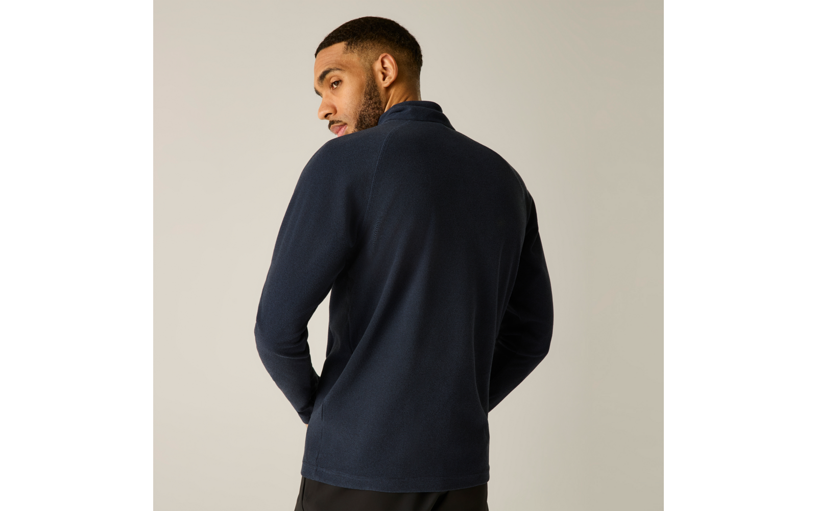 Person von hinten, Kopf nach links gedreht, trägt marineblauen Pullover und dunkle Hose, heller einfarbiger Hintergrund