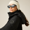 Person mit weißem, flauschigem Stirnband, schwarzer Sonnenbrille und schwarzer Kapuzen-Winterjacke; braunes Haar zurückgebunden, hellgrauer Hintergrund