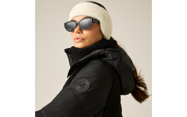 Person mit weißem, flauschigem Stirnband, schwarzer Sonnenbrille und schwarzer Kapuzen-Winterjacke; braunes Haar zurückgebunden, hellgrauer Hintergrund