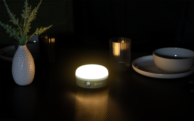 Runde, hell leuchtende Leuchte auf dunklem, leicht reflektierendem Tisch; links weiße Vase mit grünem Zweig; rechts gestapelte Teller und eine Schale; im Hintergrund zwei gerippte Gläser mit warmem Licht