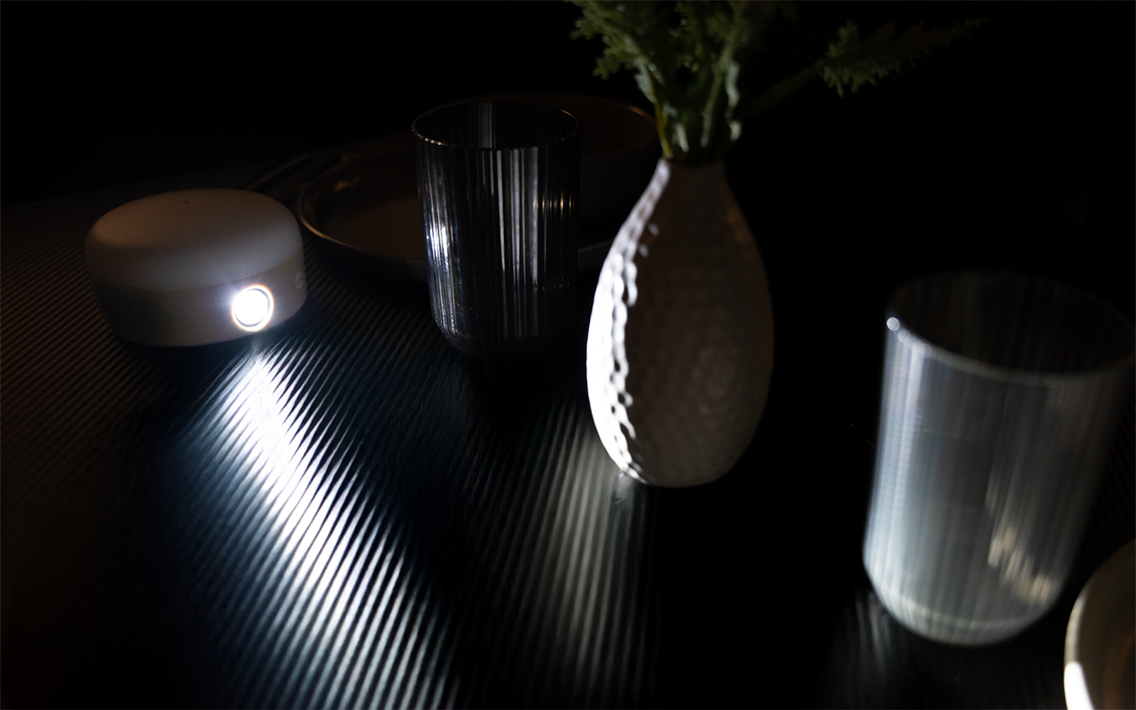 Rundes weißes Gerät mit hellem Licht links; Lichtstreifen auf geriffelter dunkler Oberfläche; weiße strukturierte Vase mit grünen Blättern in der Mitte; zwei durchsichtige geriffelte Gläser rechts und hinten