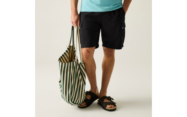 Unterkörper einer Person in schwarzer Shorts und türkisfarbenem Oberteil, mit schwarzen Sandalen und einer grün-weiß gestreiften Stofftasche in der Hand
