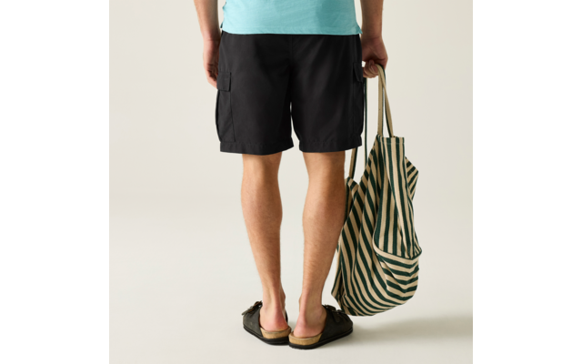 Rückansicht einer stehenden Person von der Taille abwärts, trägt hellblaues Shirt, schwarze Shorts und schwarze Sandalen, hält in der rechten Hand eine grün-weiß gestreifte Stofftasche