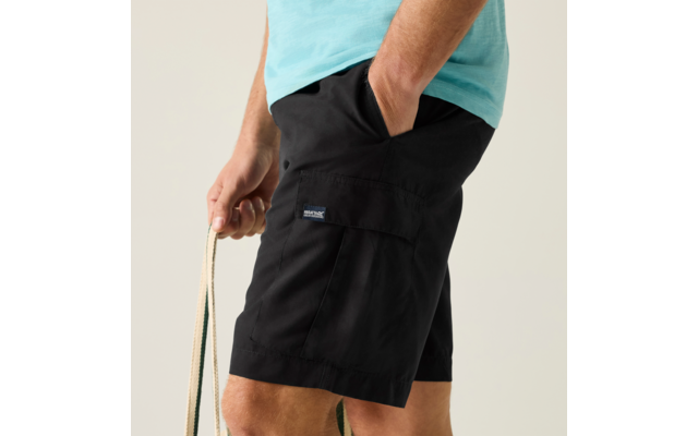 Unterkörper einer stehenden Person in türkisfarbenem T-Shirt und schwarzen Cargo-Shorts, rechte Hand in Tasche, linke Hand hält ein Seil