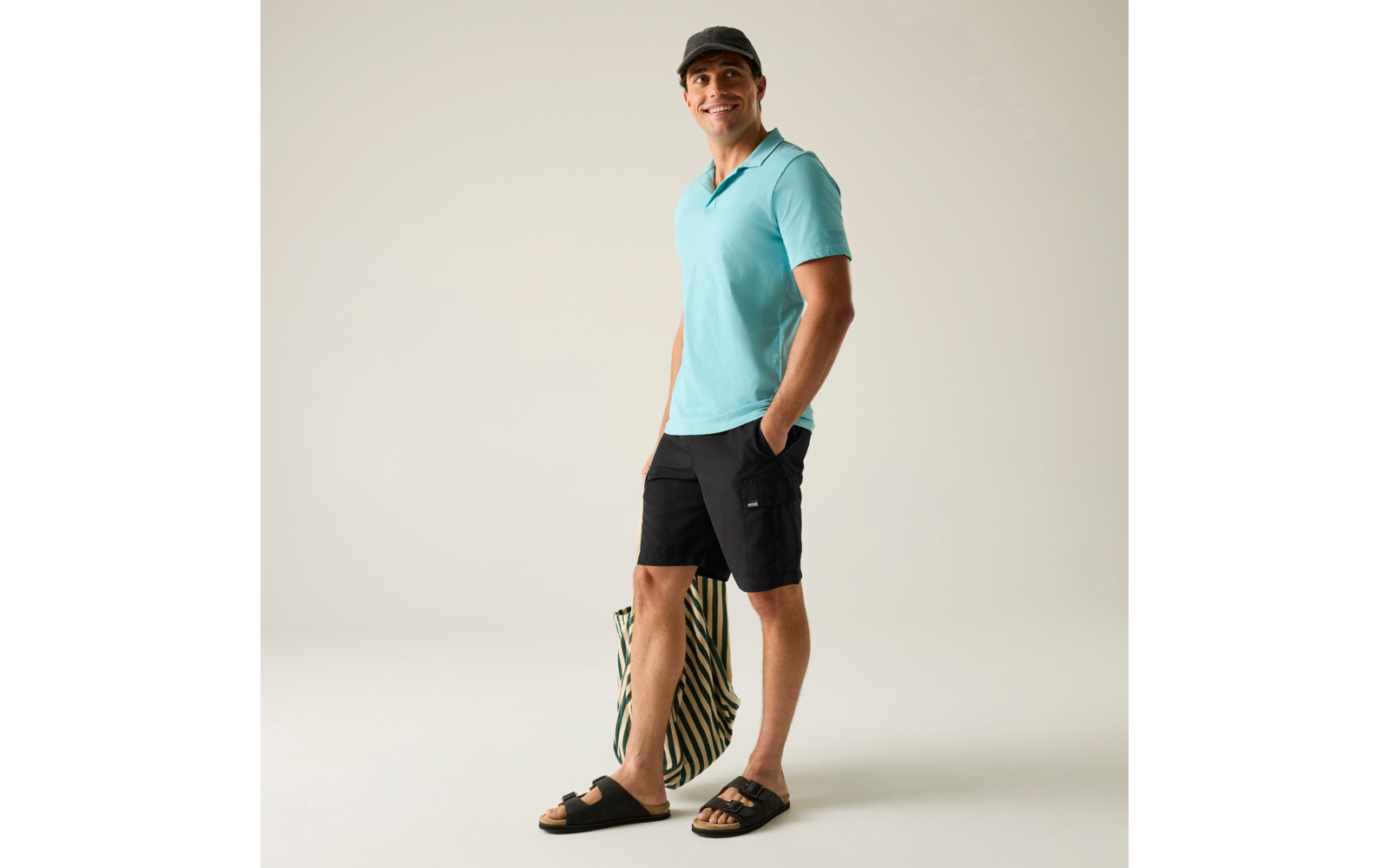 Lächelnde Person in türkisfarbenem Polo, schwarzer Kappe, schwarzen Shorts und schwarzen Sandalen, steht mit einer grün-beigen gestreiften Tasche neben den Beinen vor hellem Hintergrund