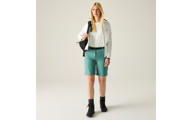 Person mit blondem, schulterlangem Haar, weiße Jacke, weißes Top, türkisfarbene knielange Shorts, schwarzer Rucksack über der rechten Schulter, schwarze Socken und schwarze Wanderschuhe, vor hellem einfarbigem Hintergrund