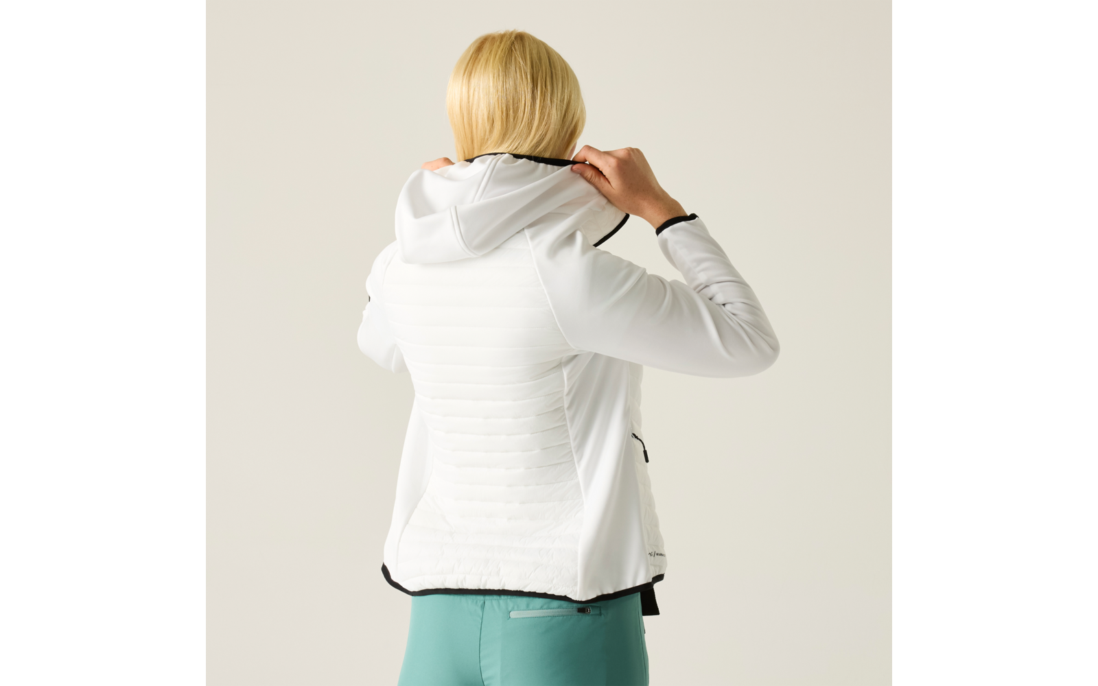 Person mit blonden Haaren von hinten, trägt weiße Kapuzenjacke und türkisfarbene Hose, hebt die Kapuze mit beiden Händen