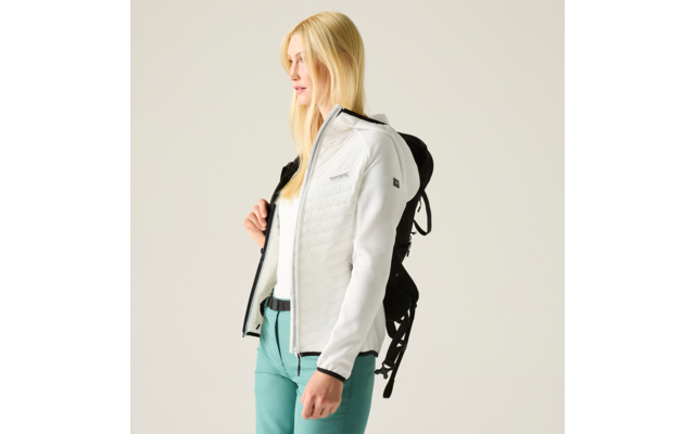 Person mit langen blonden Haaren, weiße gesteppte Jacke, türkise Hose, seitlich stehend, schwarzer Rucksack, heller Hintergrund