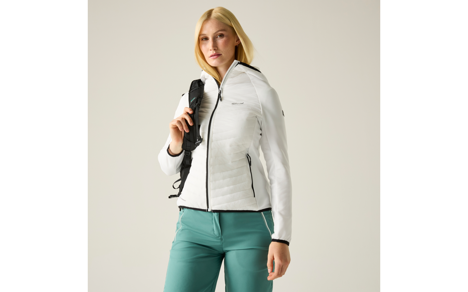 Person mit blonden schulterlangen Haaren, weißer gesteppter Jacke, grünen Hosen und Rucksackträger über einer Schulter vor hellem Hintergrund