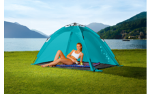 Vrouw zit in een blauwe tent op gras met een boek, bergen en een meer op de achtergrond