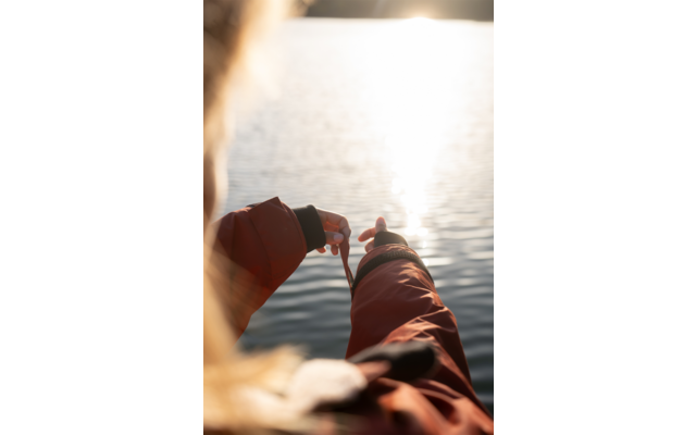 Rückenansicht einer Person in orangefarbener Jacke, die mit beiden Händen eine Schnur über ruhigem Wasser mit heller Sonnenreflexion hält