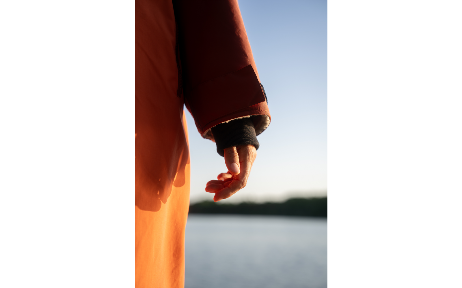 Nahaufnahme einer ausgestreckten Hand mit orangefarbener Jackenärmel vor Wasser und klarem Himmel