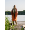 Person steht auf einem Holzsteg an einem See, trägt orangefarbene Jacke und helle Hosen, Schilf im Vordergrund, Baumreihe am gegenüberliegenden Ufer, heller Himmel
