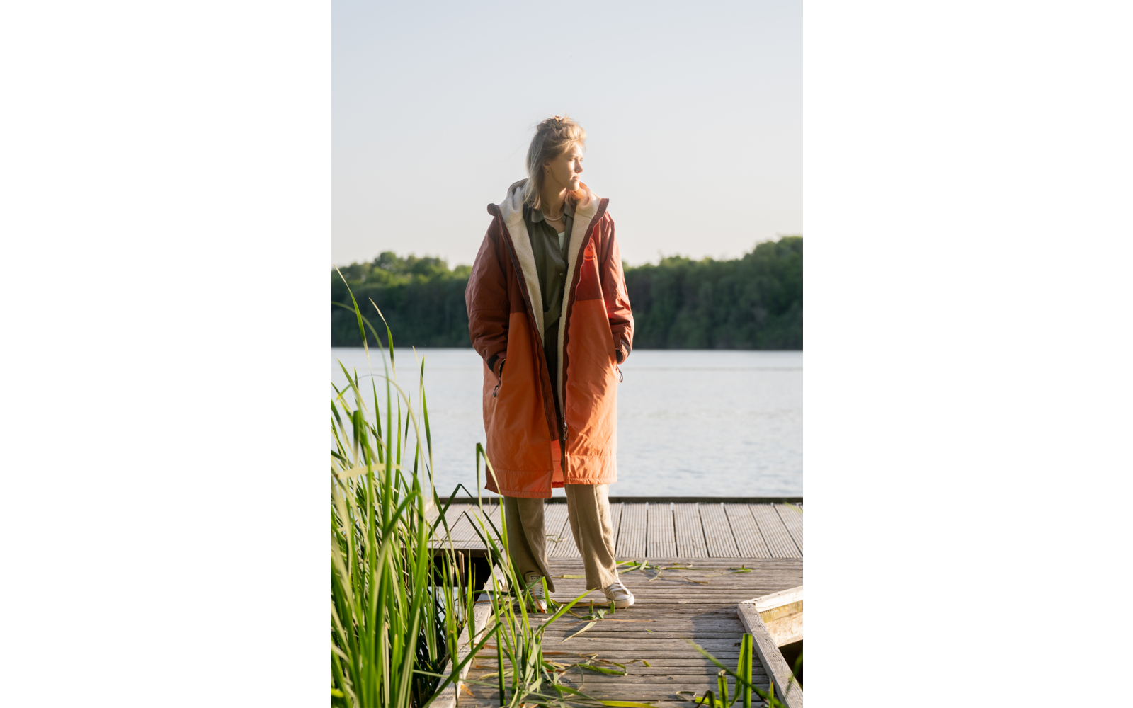 Person steht auf einem Holzsteg an einem See, trägt orangefarbene Jacke und helle Hosen, Schilf im Vordergrund, Baumreihe am gegenüberliegenden Ufer, heller Himmel