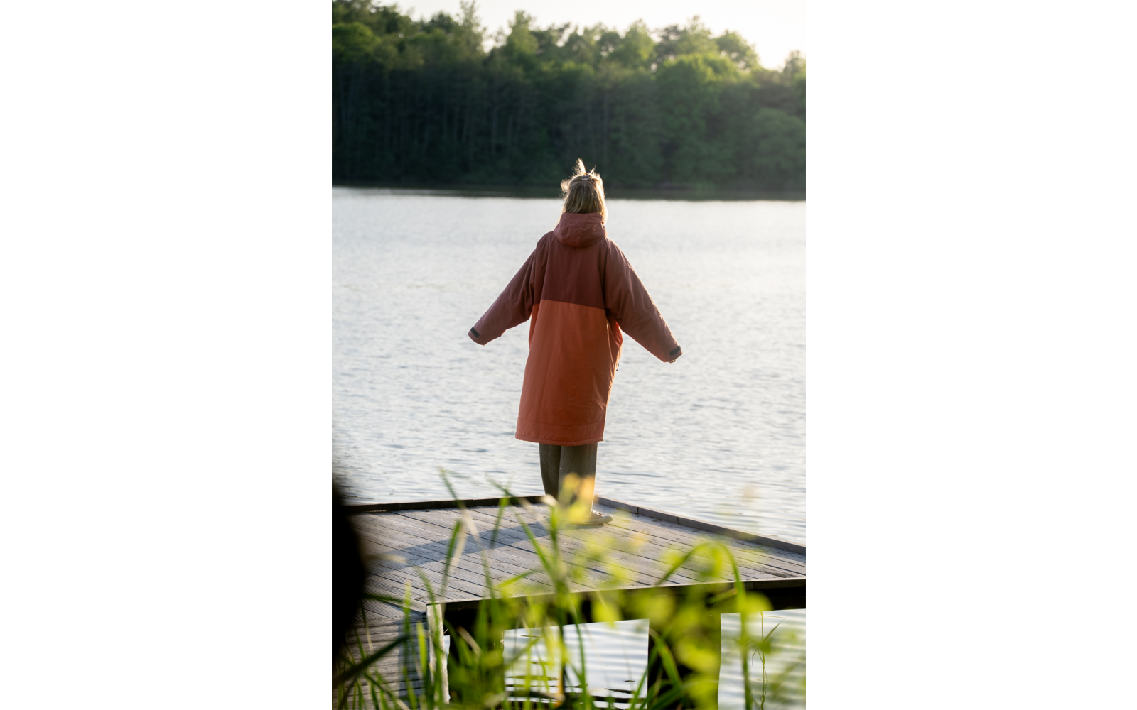 Person mit orangefarbenem Mantel steht mit dem Rücken zur Kamera auf einem Holzsteg am See; im Hintergrund Wald