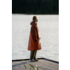 Person im Profil steht auf einem Holzsteg am See, trägt langen rostorangefarbenen Mantel und helle Schuhe, Wasser und Baumreihe im Hintergrund