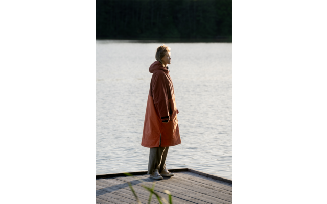 Person im Profil steht auf einem Holzsteg am See, trägt langen rostorangefarbenen Mantel und helle Schuhe, Wasser und Baumreihe im Hintergrund
