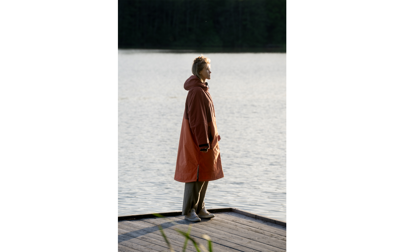 Person im Profil steht auf einem Holzsteg am See, trägt langen rostorangefarbenen Mantel und helle Schuhe, Wasser und Baumreihe im Hintergrund