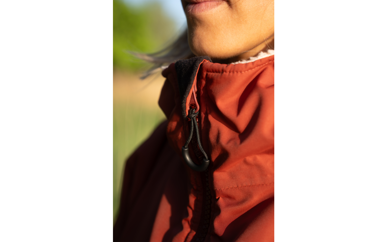 Nahaufnahme von Mund, Kinn und Hals; rote Jacke mit Reißverschluss und schwarzem Kordelzug; weißer Rüschenkragen; unscharfer grüner Hintergrund; Sonnenschein auf der Haut