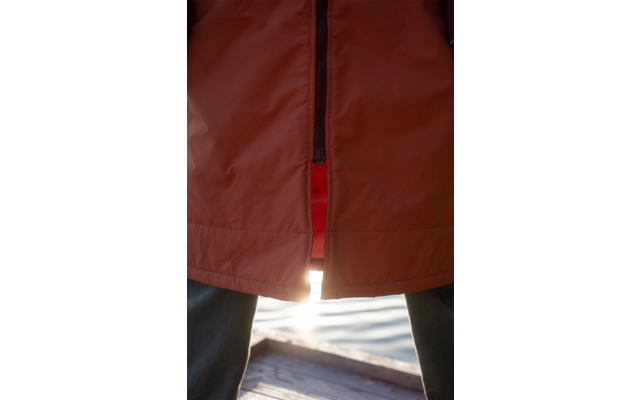 Orange Jacke mit unten leicht geöffnetem Reißverschluss, rote Innenlage sichtbar, dunkle Hosen, Holzsteg und Wasser mit Sonnenreflexion zwischen den Beinen