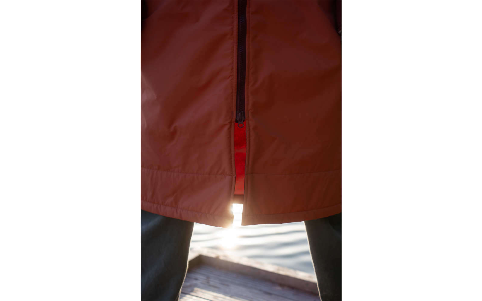 Orange Jacke mit unten leicht geöffnetem Reißverschluss, rote Innenlage sichtbar, dunkle Hosen, Holzsteg und Wasser mit Sonnenreflexion zwischen den Beinen