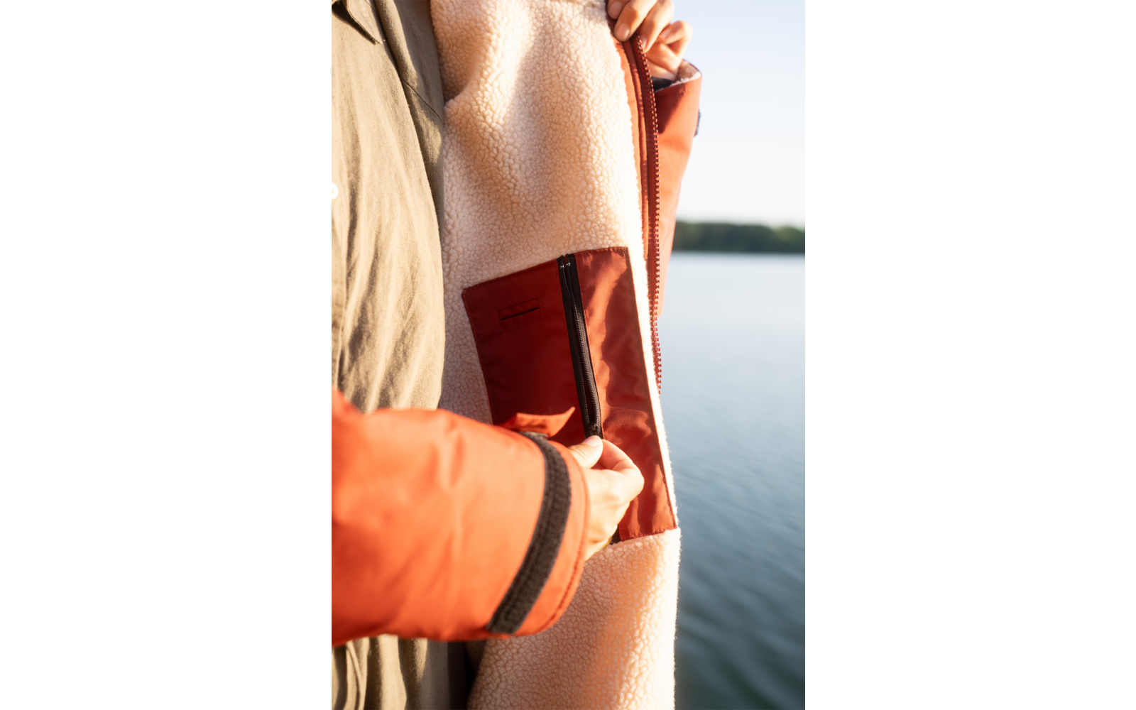 Hände an geöffneter Innentasche einer fleecegefütterten Jacke mit orangefarbenem Ärmelsaum; im Hintergrund Wasser und Ufer